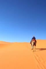 A lone figure rides a camel across expansive, orange sand dunes under a vibrant blue sky