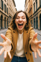 A young woman in a brown coat joyfully reaching out with open arms in a city street.