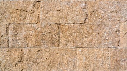 Close-up of a textured stone wall made of rectangular sandstone blocks. The rough surface features natural imperfections, earthy beige tones, and a rugged, organic appearance. Ideal for background.