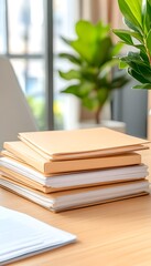 Stack of beige folders and papers on a light wood desk near a window and plant