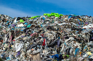 A vast accumulation of discarded clothing and fabrics fills the landfill, highlighting the issue of textile waste under bright sunlight