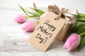 Happy Mother's Day tag attached to kraft paper gift box with tulips and ribbon on white wooden background