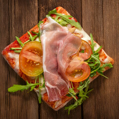 Italian pizza slice with tomato, raw ham, mozzarella and rocket on rustic wooden table. Traditional style, bright colours, simple composition, appetising dish.