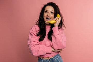 Merry young woman posing for the camera on the pink background with her favorite yellow fruit. Fun...