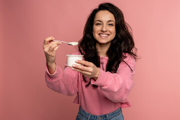 Smiling pleased dark-haired young lady eating an appetizing dairy product in front of the camera on the pink background. Healthy food concept