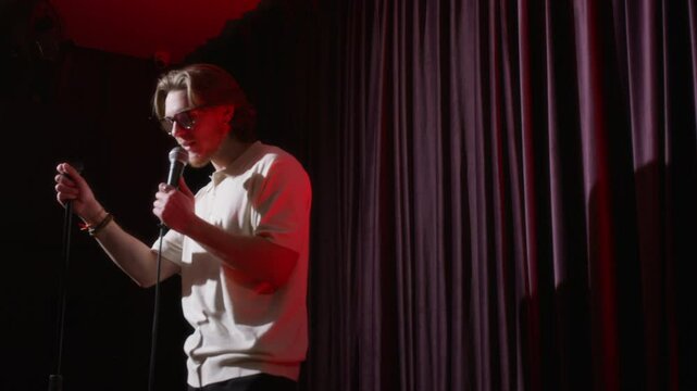 Young male stand-up artist performing on stage, talking to audience in dimly lit room with curtains