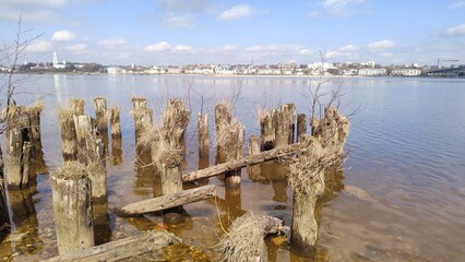 Wooden pillars from old ruined pier against backdrop of wide river and beautiful city on opposite shore. Rotten piles from pier. View of Volga River and Kostroma city