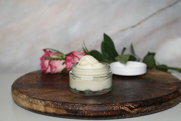 Homemade natural cream in clear glass jar on rustic dark wooden board, decorated with fresh pink roses. Organic skincare product, perfect for beauty, spa, wellness, and cosmetic themes.