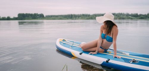 A young woman with an open swimsuit swims on a SUP board on a picturesque lake. Evening tour. A beautiful, slender girl is engaged in sap surfing on the calm water of a picturesque pond.