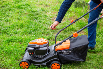 A man starts the engine of a petrol lawn mower with a starter rope. Mowing grass in the spring in a...