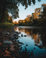 Obraz premium Serene river landscape with autumnal foliage and rocky shoreline reflecting the golden light of sunset in tranquil nature scenic view