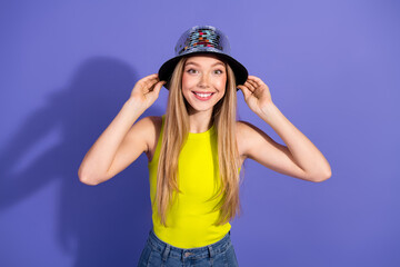 Cheerful young woman in trendy outfit adjusting shiny hat with joyful expression against vibrant purple background
