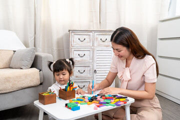 Fototapeta premium Asian young mother sitting cross-legged teaching preschool daughter sitting on floor learning creative drawing activity with colored pencils enjoying family time together at home play area.