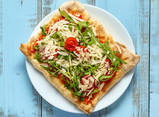 Piece of Italian pizza by the slice with tomato, rocket and Parmesan cheese on a white plate, on a shabby style light blue wooden table. Rustic style, bright colours, simple composition