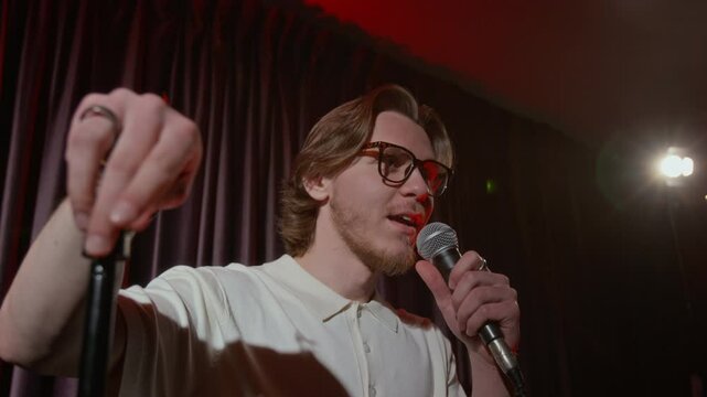 Low angle shot of young bearded man in glasses giving emotional stand-up speech on dimly lit stage