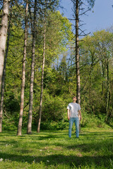 A man stands amidst a tranquil forest clearing, surrounded by tall trees and lush greenery under a blue sky, evoking peace and natural beauty.