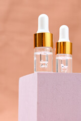 Two transparent glass serum dropper bottles with white tips and golden collars placed on a soft pink pedestal, against a peach background. Minimalist skincare branding with clean aesthetics