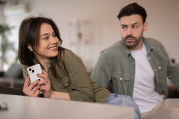 Young woman smiling with smartphone while man looks surprised in background
