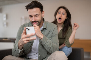 Young man focused on smartphone while surprised woman reacts in background at home
