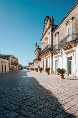 Fototapeta premium Serene street view of Noto Sicily with ornate baroque buildings under a clear blue sky casting long shadows on the cobblestone road creating a peaceful atmosphere