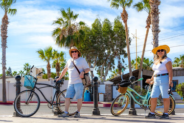 Smiling tourist couple posing next to bicycles on seafront promenade, houses and palm trees in background, shorts, hat and white blouse, sunny winter day in Loreto in Baja California Sur, Mexico