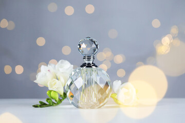 Bottle of aromatic perfume and flowers on light table against grey background with blurred lights, closeup. Bokeh effect