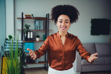 Confident businesswoman in casual office setting welcomes with an open gesture, surrounded by professional workspace
