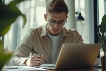 Fototapeta premium Serious and confident male businessman using laptop computer, taking notes. Work project in modern office. 25 year old male. Finance man working with documents in office. Successful business