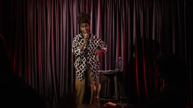 Playful African American woman giving stand-up performance on stage with microphone and purple curtains in background