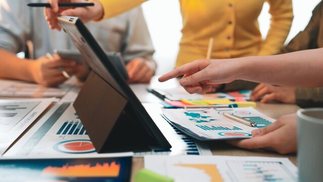 A diverse business team engages in an intense collaborative session, analyzing charts and data on a tablet, surrounded by reports and notes in a modern office environment. SACTR