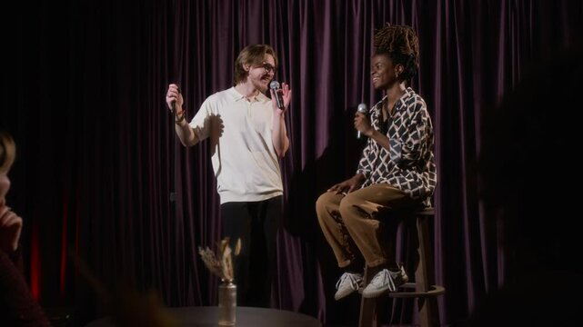Man and woman talking into microphones on stage and shaking hands during stand-up performance