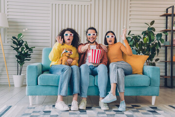 Three friends wearing 3D glasses enjoying a movie night together, sharing popcorn, in a bright and cozy living room
