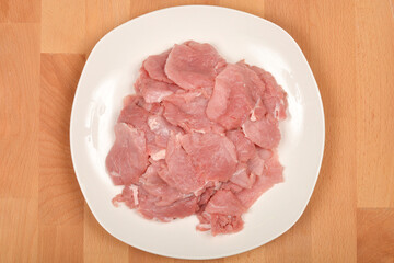 Thinly sliced raw pork tenderloin is arranged on a white plate set on a wooden surface, captured from a top-down perspective
