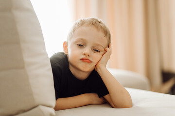 Bored child resting head on hand lying on sofa: sad little boy feeling annoyed