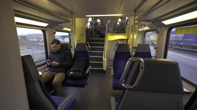 Solo traveler in winter clothing rides an empty European train through Switzerland. The man looks at his phone, surfing the internet, passing time during a long, quiet journey on a scenic route
