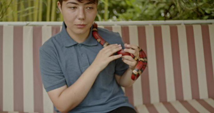 A teenage girl sits on a bench outside, playing with her pet snake.