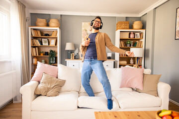 Young Man Wearing Headphones Energetically Playing Air Guitar on a Couch in a Warm Living Room