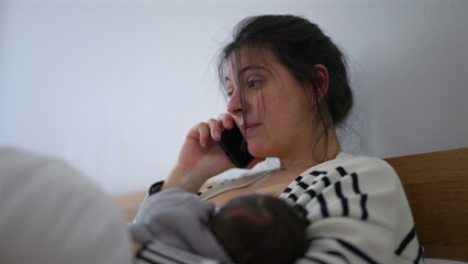Mother multitasking while her baby sleeps peacefully in her arms, speaking on the phone with a...