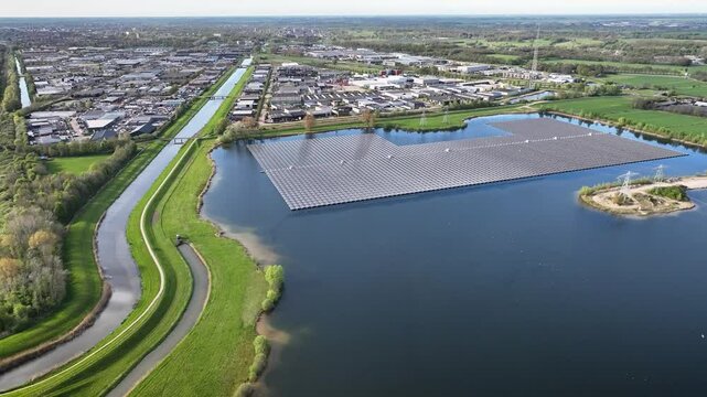 Aerial footage of large-scale floating photovoltaic system installed on a reservoir adjacent to industrial facilities with power transmission pylons on a small island.