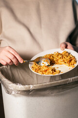 Vertical image of woman throwing away food leftovers into garbage bin trash. Scraping food with spoon. Compost from leftover food. Environmentally responsible, reducing pollution.
