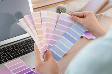 Young woman with laptop and paint palette swatches choosing lilac color near table at home, closeup