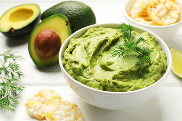 Tasty avocado dip in bowl, dill and puffed rice cakes on white wooden table, closeup