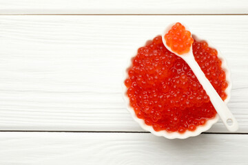 Tasty red caviar in bowl and spoon on white wooden table, top view. Space for text