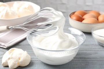 Whisk with whipped cream and other ingredients on gray wooden table, closeup. Cooking meringue cookies