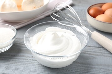 Whipped cream in bowl, sugar, eggs and whisk on gray wooden table, closeup. Cooking meringue cookies