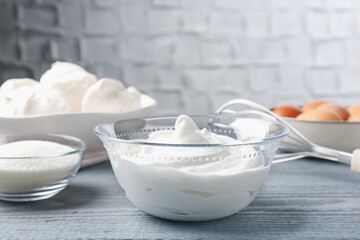 Whipped cream in bowl, sugar, eggs and whisk on gray wooden table, closeup. Cooking meringue cookies