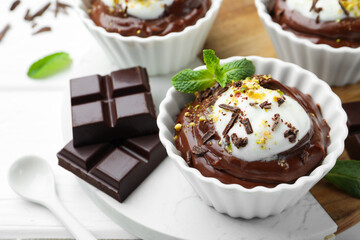 Tasty pudding with whipped cream in bowls and pieces of chocolate on white wooden table, closeup