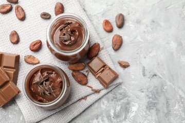 Tasty pudding in glass jars, pieces of chocolate and cocoa beans on light grey table, flat lay. Space for text