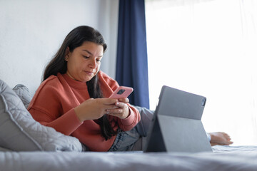 Latina woman using smartphone and digital tablet on bed at home