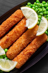 Homemade breaded cod fish fingers with garden peas and sliced lemon in a black stoneware dish. On a black wood background.